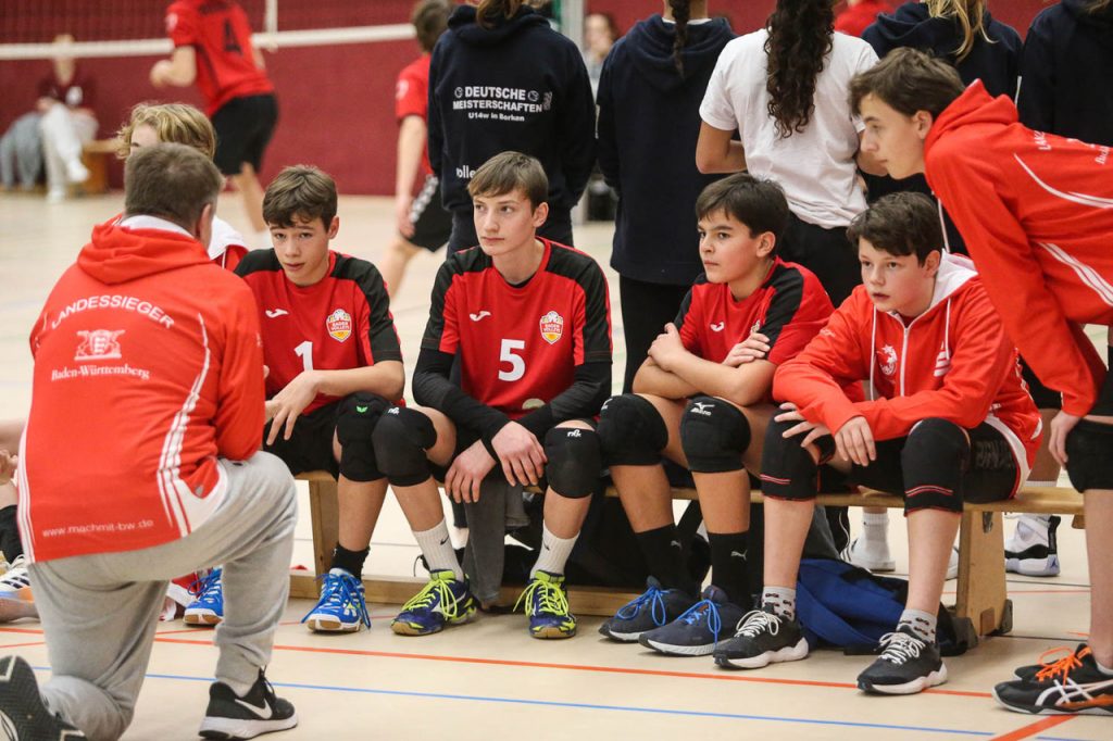 Fünf junge Volleyballer in roten Trikots und schwarzen Hosen sitzen auf einer Bank in der Halle und hören dem Trainer zu, der vor ihnen kniet.