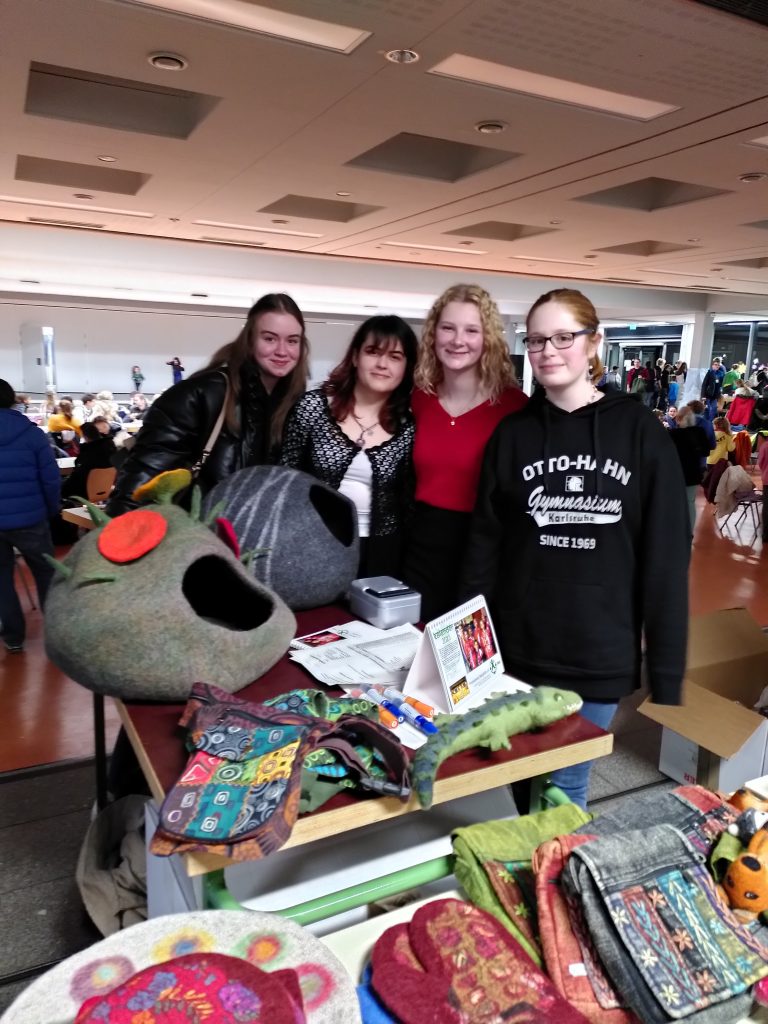 Vier Schülerinnen stehen vor dem Verkaufstisch des "Nepal-Projektes". Im Vordergrund sind bunte Taschen, Untersetzer aus Filz und kleine Tierfiguren zu sehen. Im Hintergrund der gut besuchte Weihnachtsbasar in der Aula des Otto-Hahn-Gymnasiums.