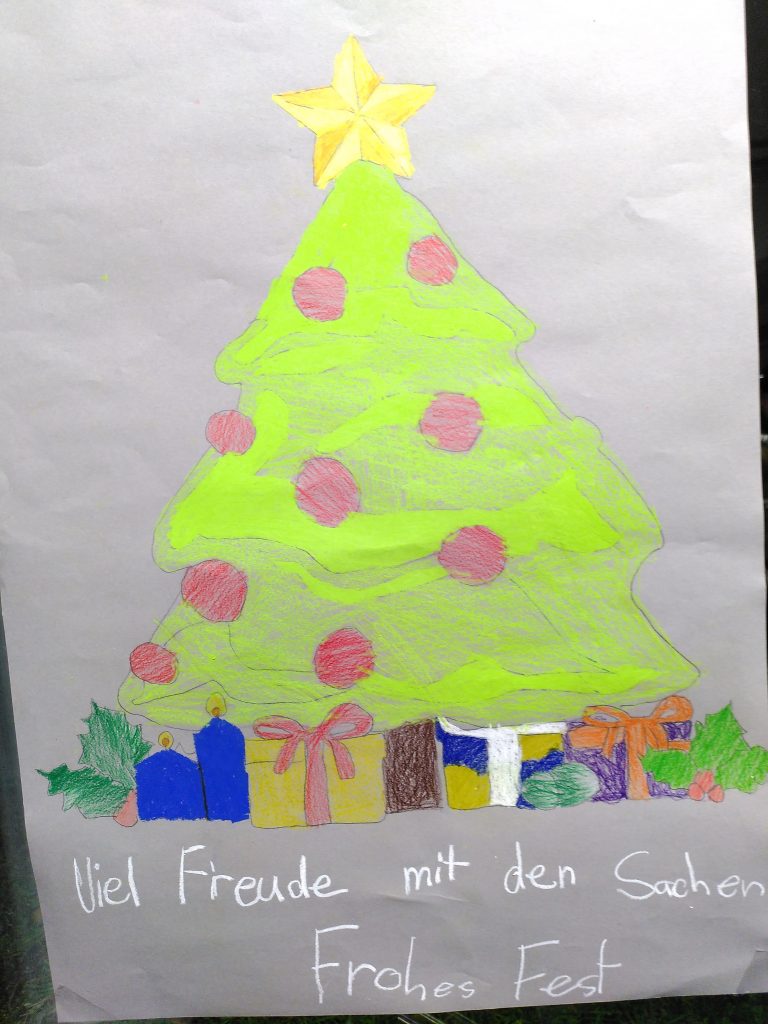 "Viel Freude mit den Sachen" steht in weißer Schrift auf dem mit einem Weihnachtsbaum und darunterliegenden Geschenken bemalten Plakat.