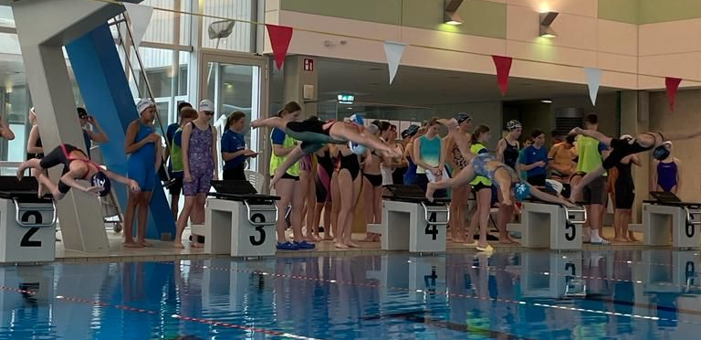 Schwimmhalle mit Blick auf die Startblöcke und dahinter stehende SchülerInnen. Vier SportlerInnen sind in der Luft kurz nach dem Absprung zu sehen.