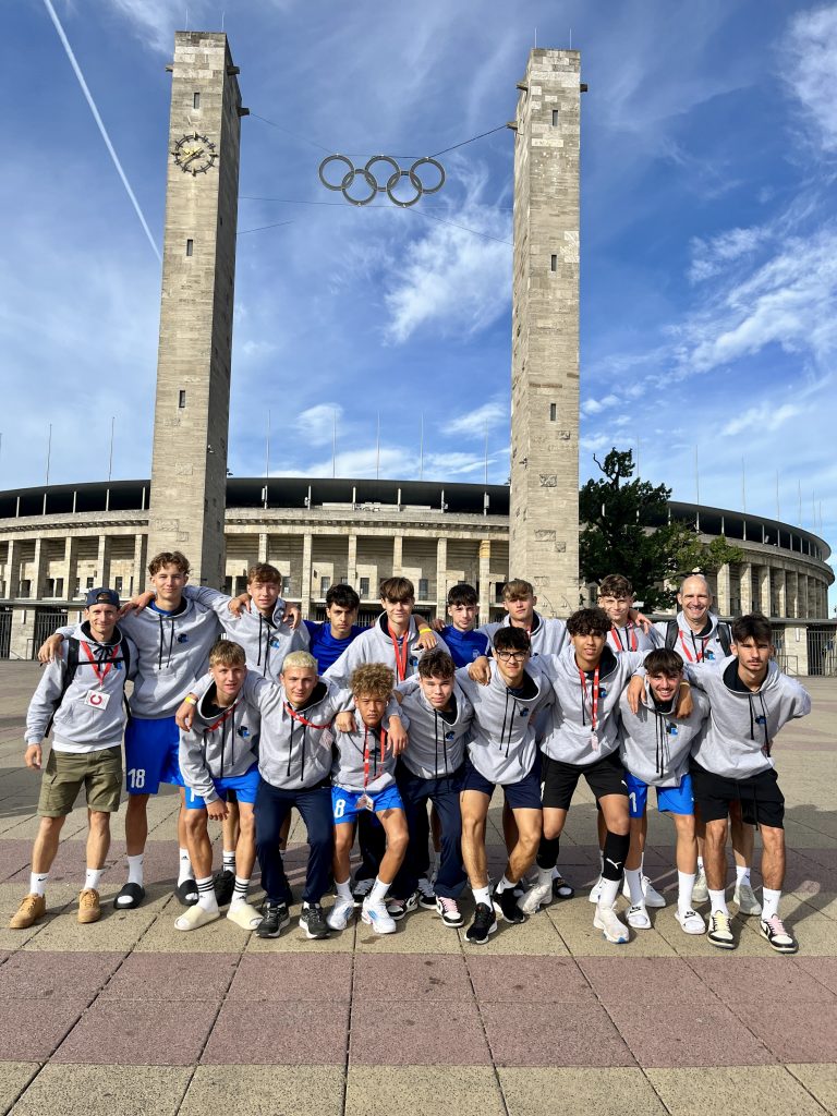 Die in graue OHG Pullover gekleideten OHG-Fußballer mit ihren Trainern J. Mudrak und J. Faustmann in Berlin. Sonniger Himmel im Hintergrund die olympischen Ringe.