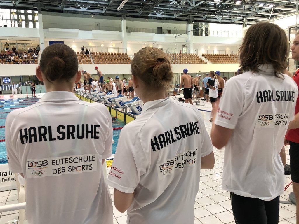 Man sieht drei SchülerInnen in der Schwimmhalle von hinten. Trikotaufschrift Karlsruhe Eliteschule des Sports.